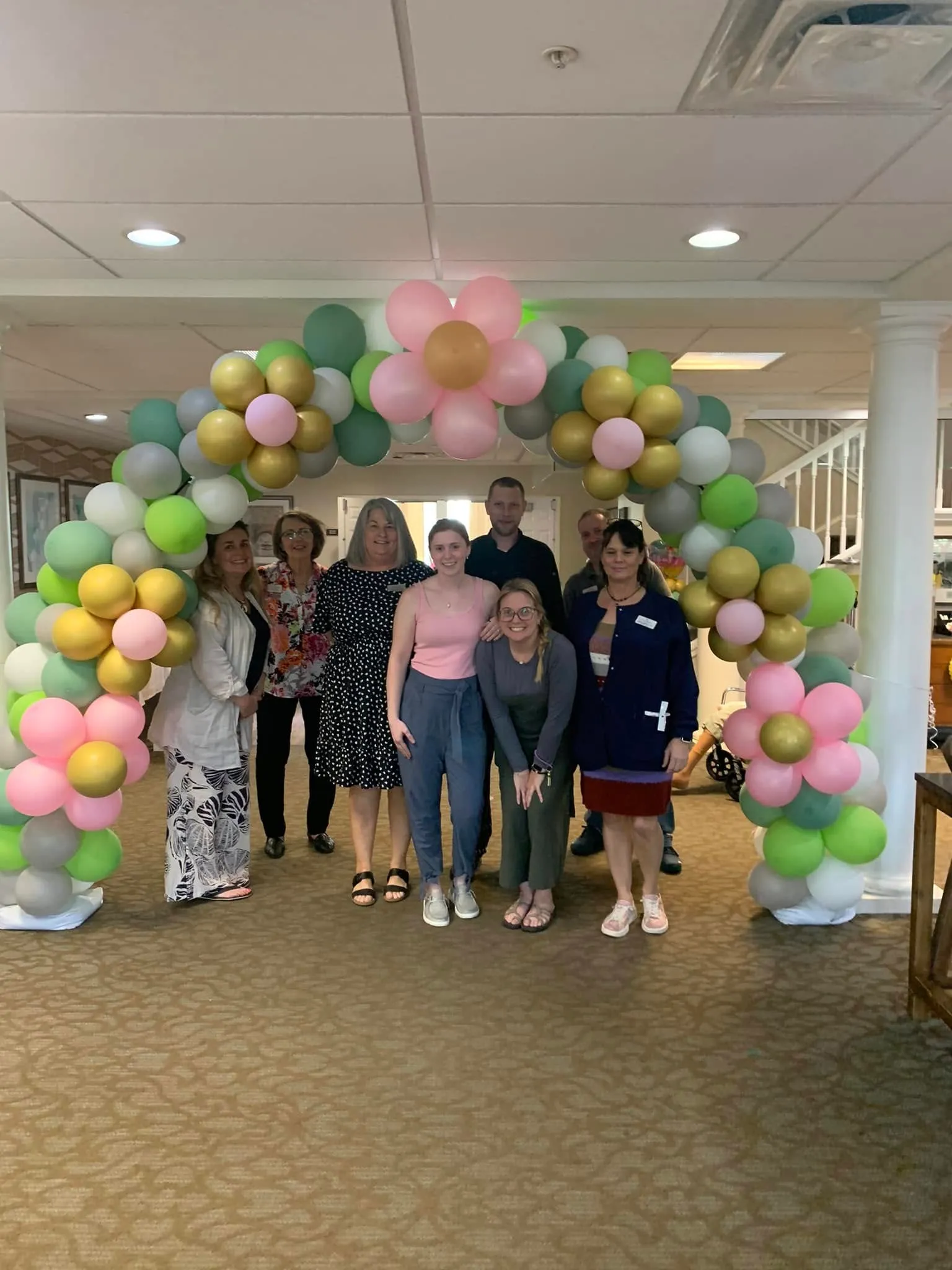 Arch of sage, lime, gray and white balloons with flowers made of pink and gold balloons, with a group of people underneath