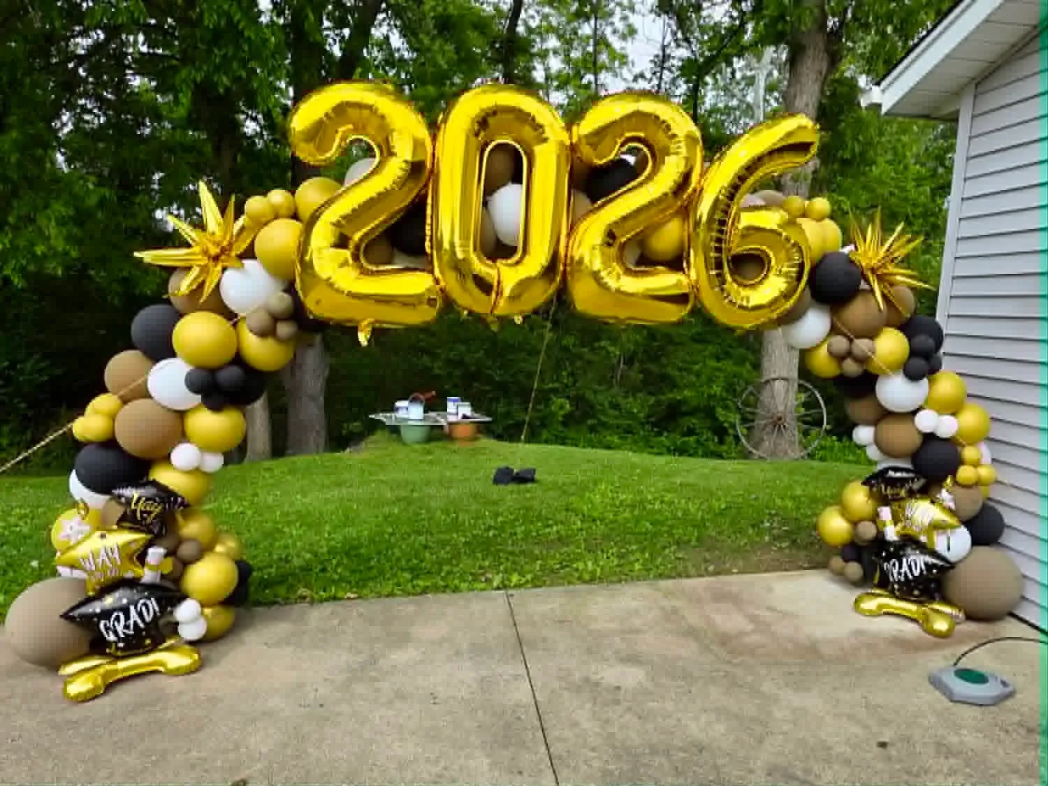 Outdoor balloon arch in gold, black, white and taupe topped with giant gold 2026 number balloons and grad foils at the base