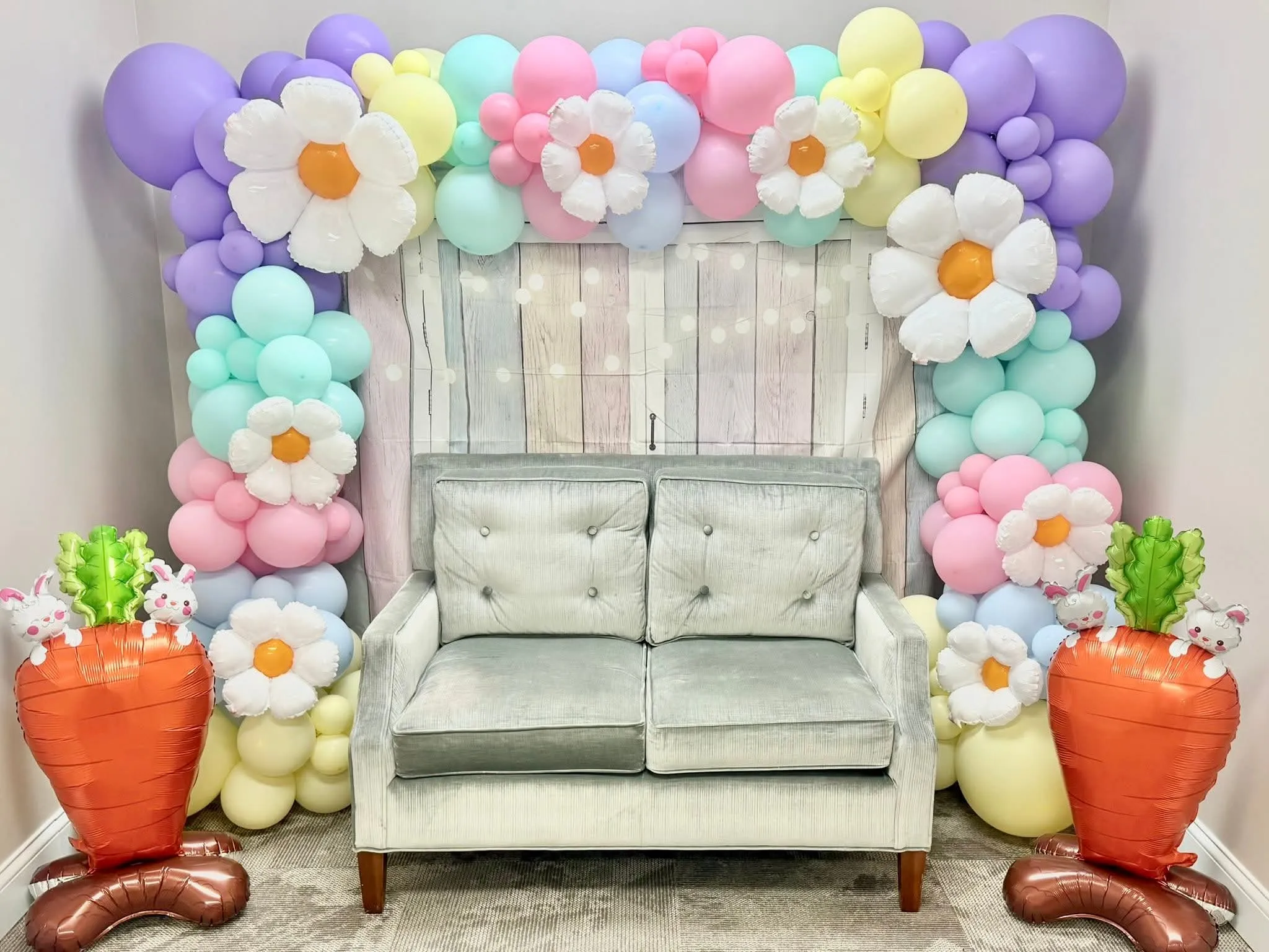 Pastel balloon garland with daisy balloons around a wooden barn backdrop, a loveseat and carrot balloons for an Easter photo booth