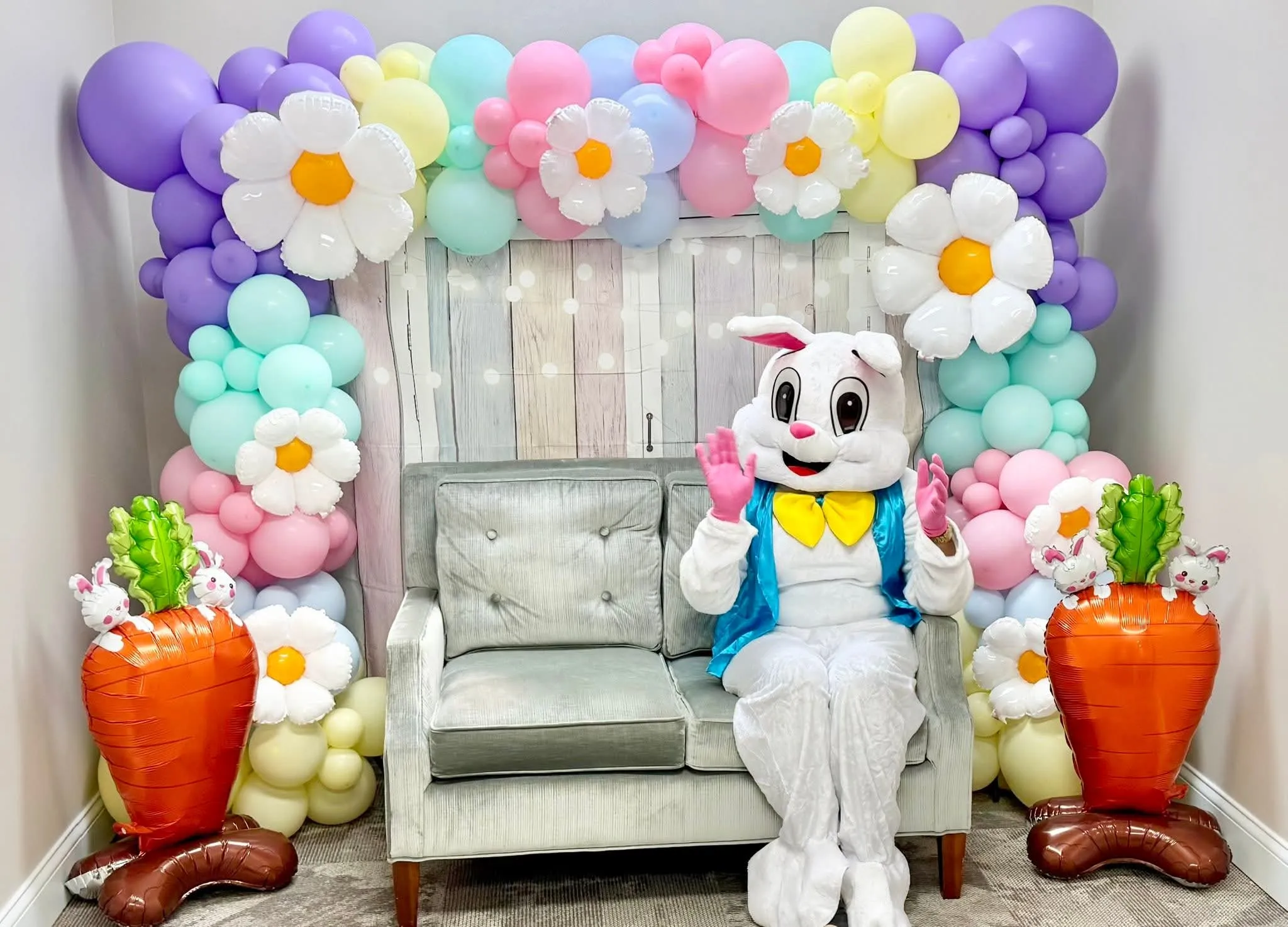 Easter Bunny waving from a loveseat inside a pastel daisy balloon garland with carrot balloons on each side