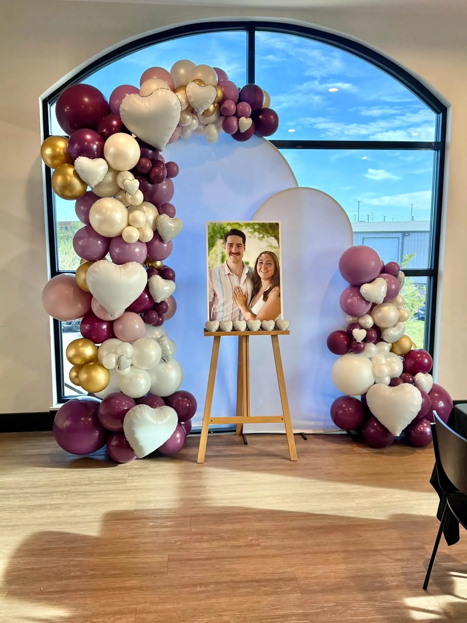Burgundy, mauve, pearl and gold balloon arch with white heart balloons framing an engagement photo on an easel