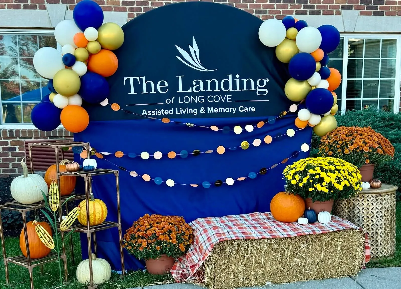 Navy, gold, orange and white balloon clusters on a branded sign backdrop with pumpkins, mums and a hay bale for a fall festival