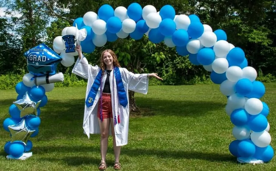 Outdoor blue and white balloon arch with a graduation cap balloon and silver stars, with a graduate in a white gown underneath