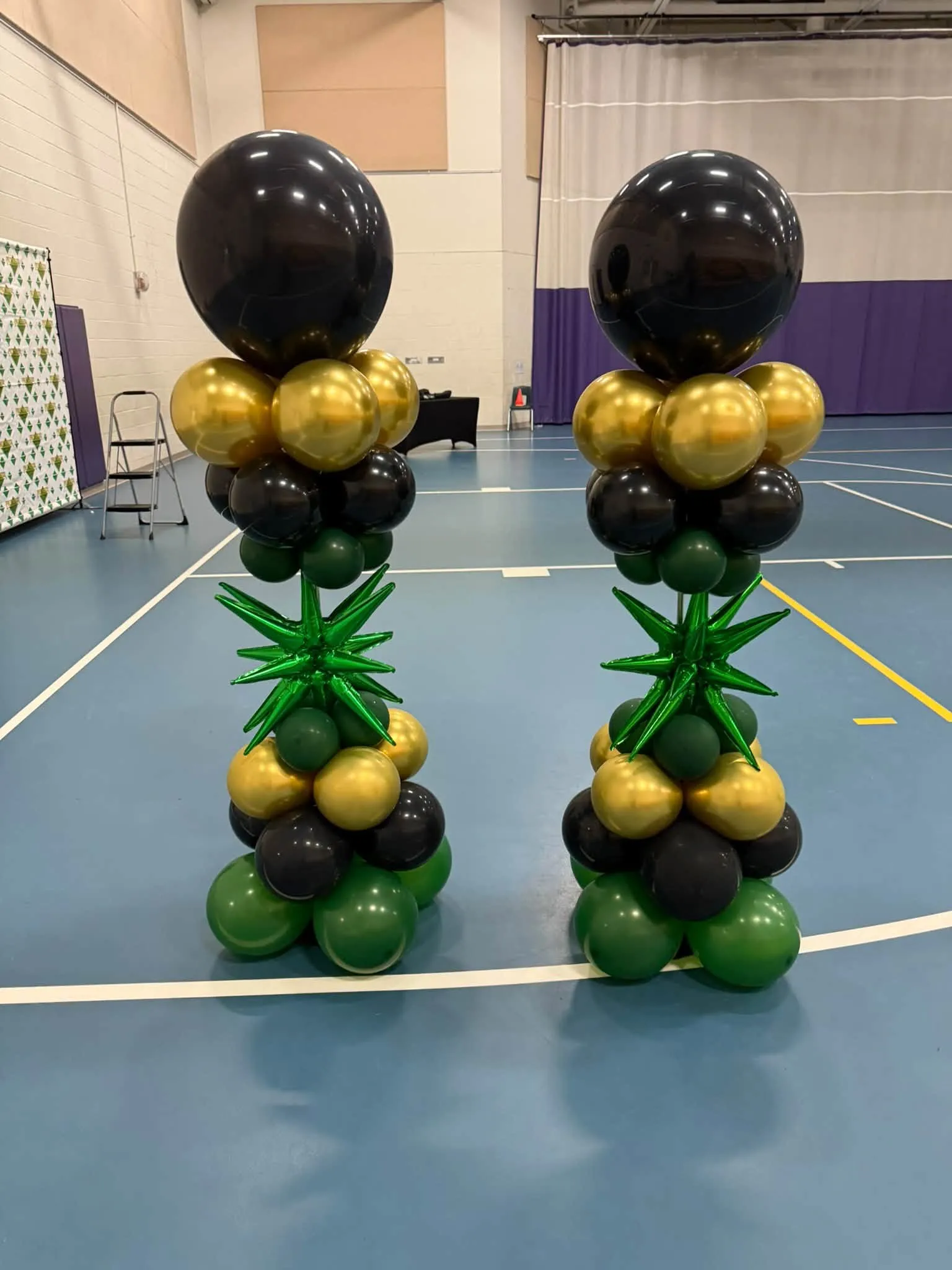 Two balloon columns of black, gold and green with green starbursts, topped with large black balloons, in a gym