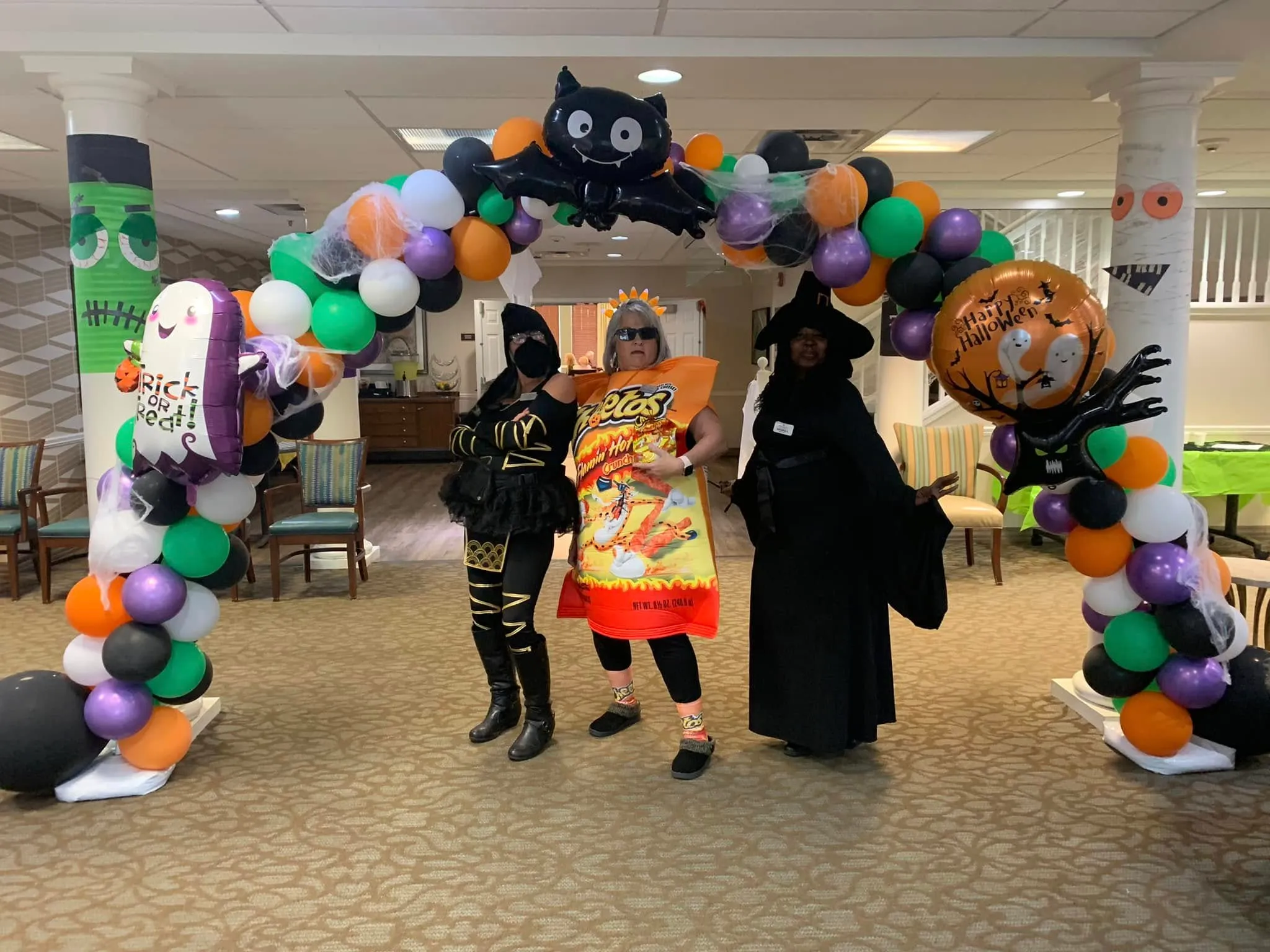 Halloween balloon arch in orange, purple, green, black and white with a black bat foil, a ghost and a Happy Halloween balloon, with three people in costume underneath