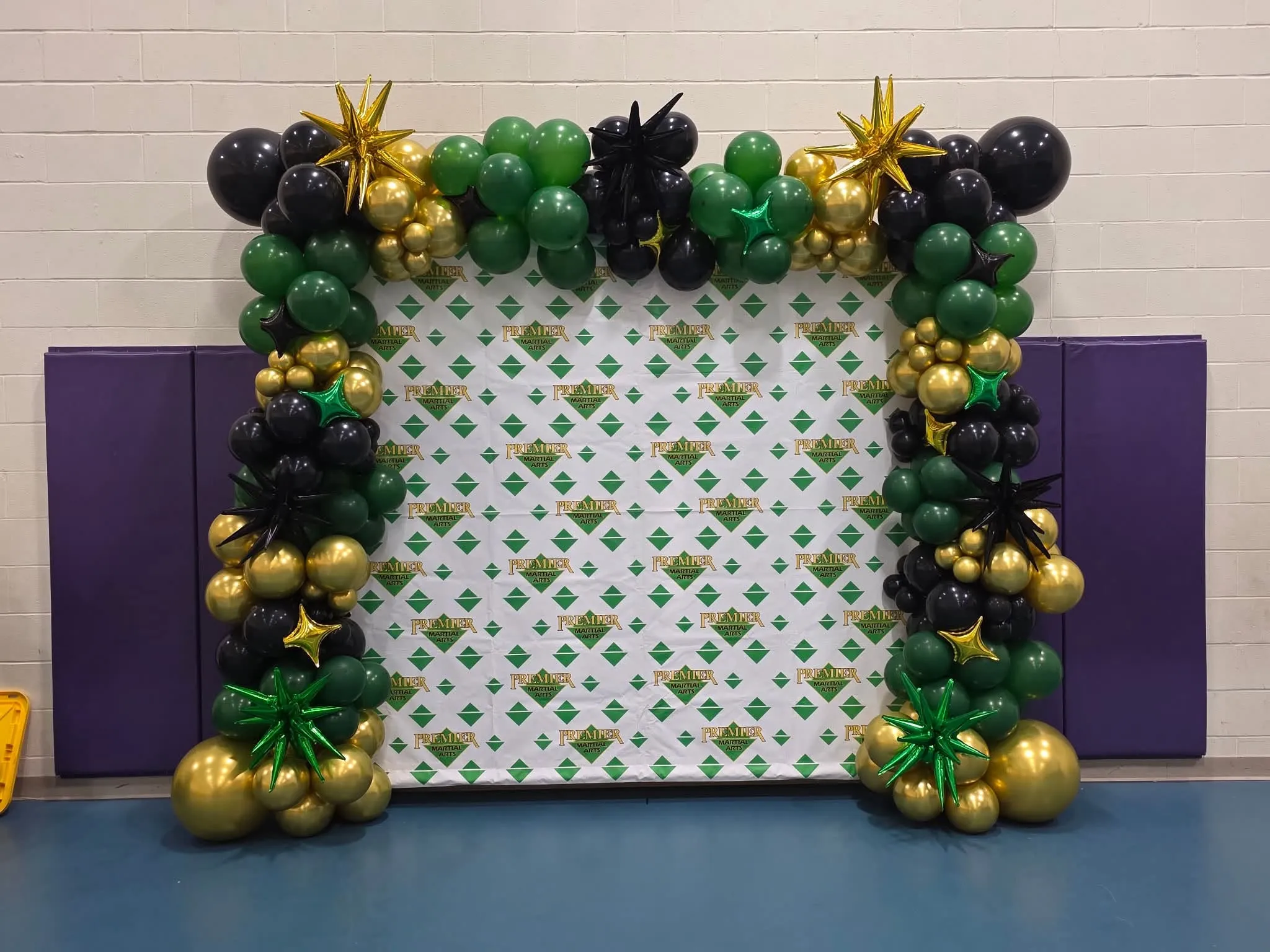 Green, black and gold balloon frame with starbursts and star foils around a logo step-and-repeat backdrop