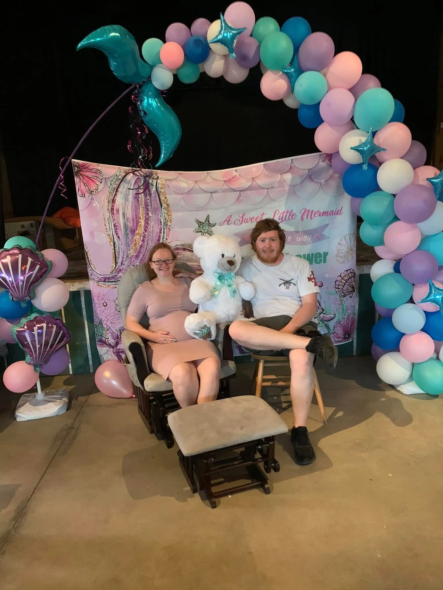 Organic arch of pink, lavender, teal, blue and white balloons with a teal mermaid tail and star foils, beside a column with purple shell balloons, framing a mermaid baby shower backdrop with the parents-to-be seated in front