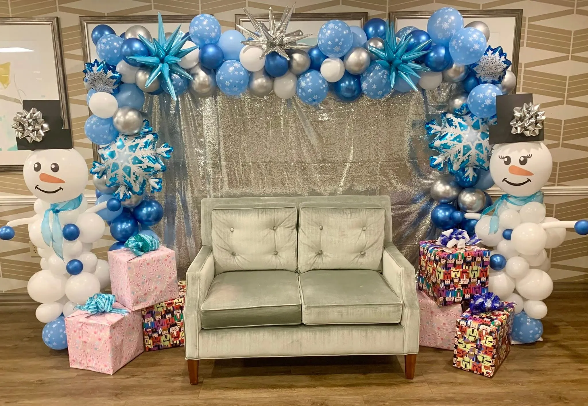 Blue, white and silver balloon garland with snowflake and starburst balloons, balloon snowmen and wrapped gifts around a loveseat